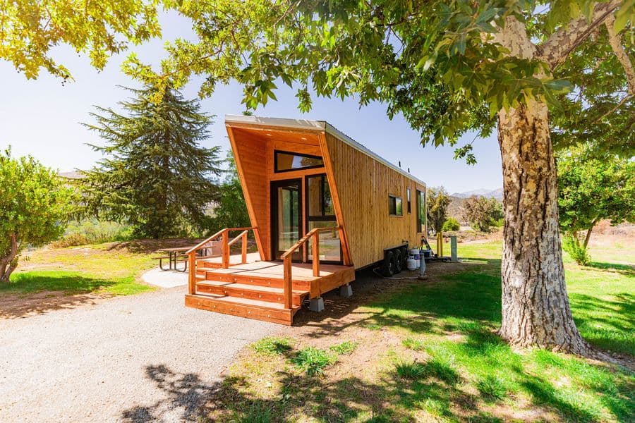 Cyrus, a wood-clad tiny home on wheels with a deck and wooden steps, under a large shade tree on a green lawn