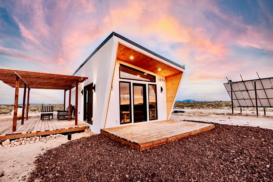 Luna, a white angular tiny home with a pergola deck and solar panels, under a pink sunset sky